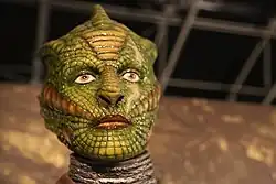 A sculpt of the head of a reptilian humanoid. Its face resembles that of a human, but is green and has lizard-like skin.