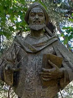 Chest-length metal statue of bearded man in robes and turban holding a book and a cubic model.