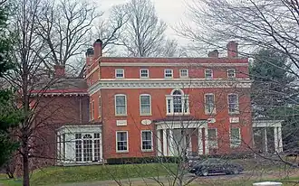 A large two-story flat-roofed rectangular brick building with four corner chimneys classically inspired ornamentation. There is a large full-height wing on the left and a small white wooden porch on the right. There are bare trees and a car parked in the front driveway.