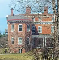The same house seen from the side with the large wing, also in springtime, illuminated by sun from the left, with some bare branches and bushes partially obscuring the view