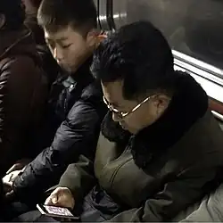 An older man is sitting down on a train, staring down while playing a tile-matching mobile game. A child sitting next to him is also looking over at his phone.