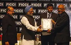 Four men in formal attire are standing on a stage in front of a black background with white text on it. The two men in the middle are both grabbing an award plaque.
