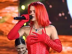 A woman with red hair is singing to a microphone with her eyes closed. She is wearing a red top and red gloves.
