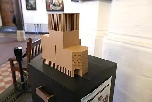 Building made up of rectangular prisms, the center one very tall, and two smaller ones surrounding it on the left and behind; the right side features a rectangular prism merged to a half-hexagonal prism of the same height. Small, frequent slit windows, a single tall vertical window and a slit arcade along the fourth side of the core, built of wood.