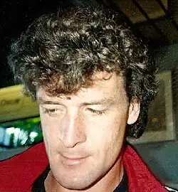 Colour photo of Mark Hughes. He is wearing a red collared jacket and is looking down and to the right