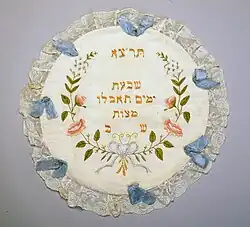 A white circle of fabric trimmed with lace, blue ribbons, and embroidery, including embroidered Hebrew letters
