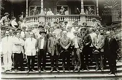 A crown of approximately 40 men standing on a set of steps in front of stone balcony