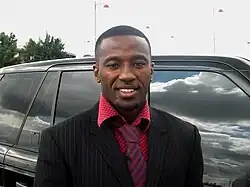 A smiling black man dressed in suit and tie stands by a large shiny car
