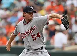 A man in a gray baseball uniform with "WASHINGTON 54" on the chest, a dark baseball cap, and a baseball glove on his left hand pitches a baseball with his right hand.