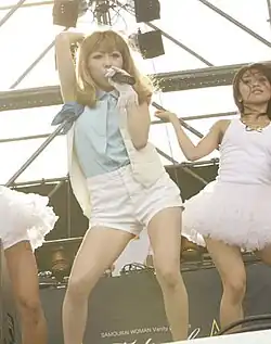 A woman in a blue shirt and white shorts sings into a microphone, flanked by two other people dancing.