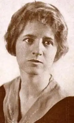 A young white woman with blond hair, wearing a dark garment with a scarf effect at the neckline