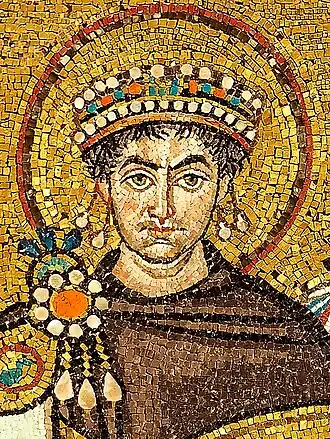A close up photograph of a mosaic of a person painted on a golden coloured background. The person has a dark brown cloth covering up his upper body till the neck, three jewels on his right shoulder, a crown with two pendants each hanging from both ends and an aura like circle around his head