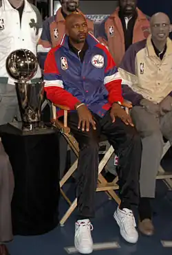 An older African-American man, wearing a black shirt and blue-red jacket with the logo NBA on it, is sitting on a chair while posing for a photo. He is surrounded by four other men, while a gold trophy is placed on his right.