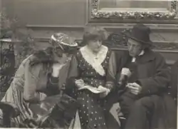 Black and white photograph showing three people on a film set, with a man in a hat sitting with two women