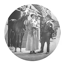 A woman in a long skirt and jockey's cap holding the reins a horse on her right with a man in a suit standing to her left.