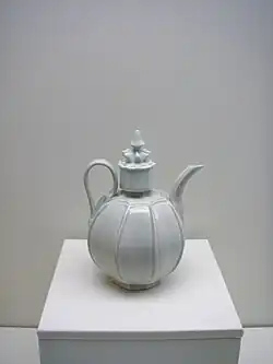 A white teapot with an almost perfectly spherical body and a large, cylindrical cap in the center which is topped with a small crown shaped embellishment. Several vertical lines are glazed into the body of the pot.