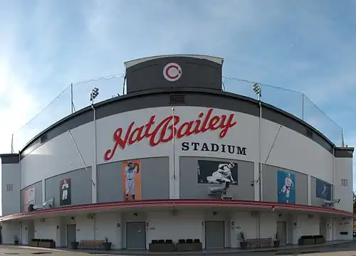A photo taken on the outside of a white stadium with wording in a semi-cursive font "Nat Bailey". Below it is wording in a Times New Roman font "Stadium". Below it are six dark grey rectangles, each having a photo of a man.