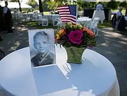 A color image showing a black-and-white photo of a young boy. The picture stands on a small round table beside a vase of flowers containing a U.S. flag.
