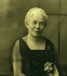 B&W photo of an elderly woman wearing a dark short-sleeved dress with square neckline.