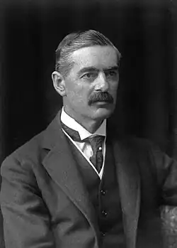 A black and white mid-length photograph of Neville Chamberlain, a man wearing a dark waistcoat and with a grey moustache, looking to the left.