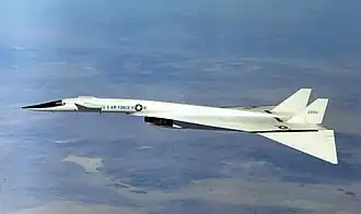 White delta-wing aircraft overflying mountains. The front of the fuselage features canard wings, and the wing tips are dropped.