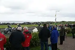 Spectators standing in the front garden of a house watching the motorcycle racing.