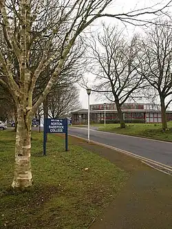 Building with multiple glass windows seen at the end of a tree lined road. Sign says welcome to Norton Radstock College