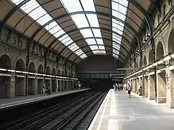 A railway station with side platforms either side of two tracks that disappear into darkness under a painted steel bridge like structure topped with a brick wall, covered by a partially glazed barrel roof.