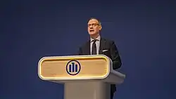 Oliver Bäte talking and standing behind a white podium with light wodden look and the Allianz logo