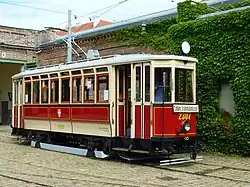 An antique ivory and white single streetcar in front of a brick building. The car has the crest of Vienna on its side, and the title "Erdberg Straßenbahnmuseum" on its front. The tram is empty except for a driver.
