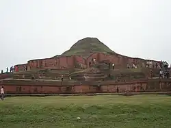 Ruins of a structure of red stone now resembling a small hill or mound.