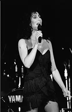Cher sings on stage in a black fringed mini dress with spaghetti straps, her dark curly hair framing her face. A drum set is visible in the background.