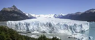 The end of a glacier, which looks like a wall of ice. Blue water filled with snow and ice is at the bottom of the cliff.