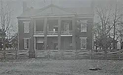 A black and white photograph of the front of an old two story brick building with a large porch and portico behind a wooden fence. A man stands in the doorway, a small painted sign advertising a lawyer's services is hung near the front door, and a gazebo is located to the right of the building.