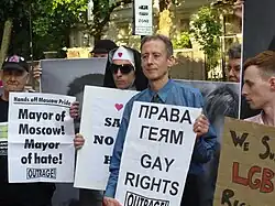 A group of people protest by holding up different signs; Peter Tatchell holds a bilingual Russian–English sign reading "GAY RIGHTS", "Outrage!"