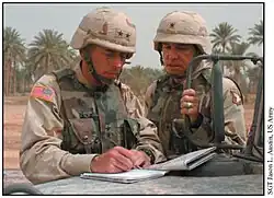two men wearing three color desert combat uniforms, both with general insignia, look at a map and notebook atop a hood of a HMMWV.