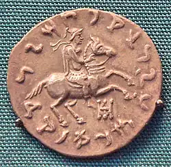 Another coin of Philoxenus. Reverse, showing the king in armour, wearing a crested Boeotian helmet, diadem, chlamys, and boots, while riding a galloping horse. He is also making a blessing gesture with his right hand.