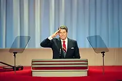 A man, wearing a suit, saluting while standing between two transparent, glass teleprompters