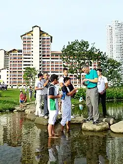 The Prime Minister is standing near the river surrounded by schoolchildren, showing him a chemistry experiment.