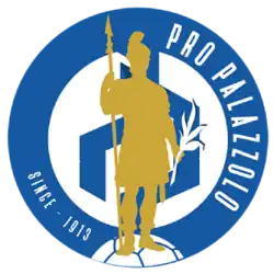A blue circle with the text "Pro Palozzolo" "Since 1913" inside of the circle. Further into the circle is another white circle containing a blue design in the middle. There is a golden silhouette of a Roman soldier in front of everything.