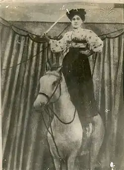 Photograph of a Gurian woman, dressed in a Victorian-style skirt and blouse, standing upon the back of a horse, with a raised sword in her right hand and her left hand on her hip.