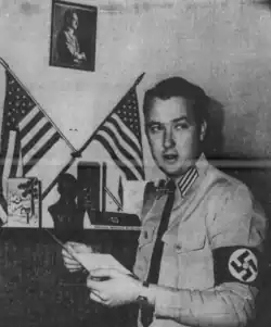 Black and white photo of Forbes holding a sheet of something and wearing a Nazi armband. Behind him there are American flags and a framed portrait of Adolf Hitler