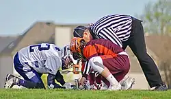 Two men wearing helmets, short-sleeved jerseys, shorts and gloves, one in a maroon and orange color scheme and the other in blue and white, kneel and crouch on grass as a man wearing a black and white vertically-striped long-sleeved shirt and long black pants with a black cap leans from behind them and places a small ball between the mesh heads of two sticks they hold on the ground between them.
