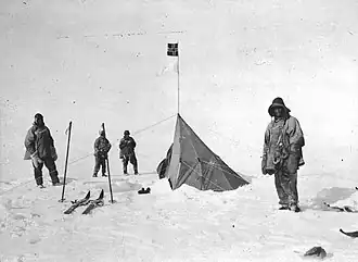 Four figures in heavy clothing stand near a pointed tent on which a small square flag is flying. The surrounding ground is ice-covered. Ski and ski poles are shown on the left.