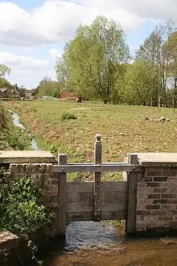 A small canal running through a field where a sluice is slightly raised with trees and a small farm in the background