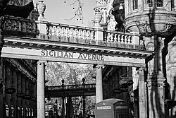 A black and white photograph detailing a colonnade of white Ionic columns with entablature. Capital letters on the entablature spell the words Sicilian Avenue