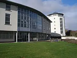 A modern 3 story building with a prominent frontage of numerous windows and constructed from a white material curves gently away from a green lawn in the foreground. In the background there is a tall white tower of a similar construction.