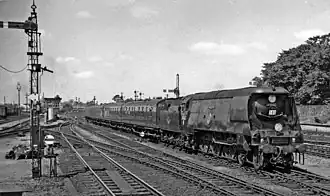 Side-and-front view of a large 4-6-2 steam locomotive with a tender, hauling a passenger train. The locomotive boiler is hidden by a casing of flat metal side sheets.