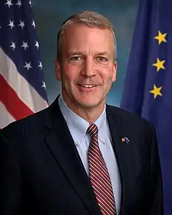Dan Sullivan's official U.S. Senate portrait as a new Senator taken in 2015, after his swearing in post-victory in 2014. The background has the American and Alaskan flags, and he is facing forwards with his body facing rightwards (towards his left hand side).