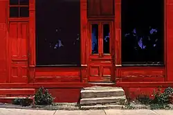 oil painting of a 200-year-old building with 2 large pane-glass windows and 2 doors, steps, weeds—painted deep red with black windows mirroring blue sky